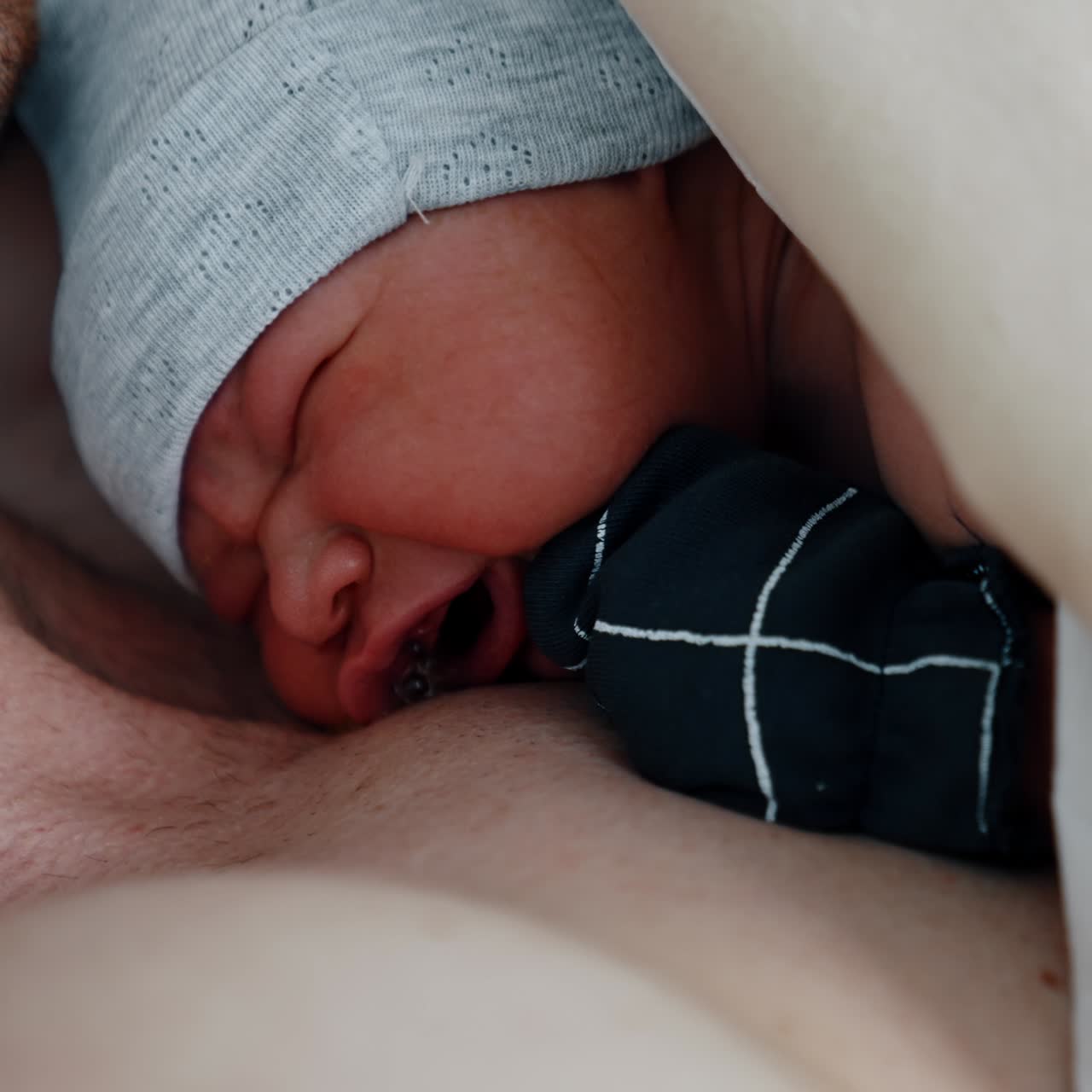 Tiny newborn is crying while lying on the chest of his dad. Baby opens eyes for the first time after being born. Close up