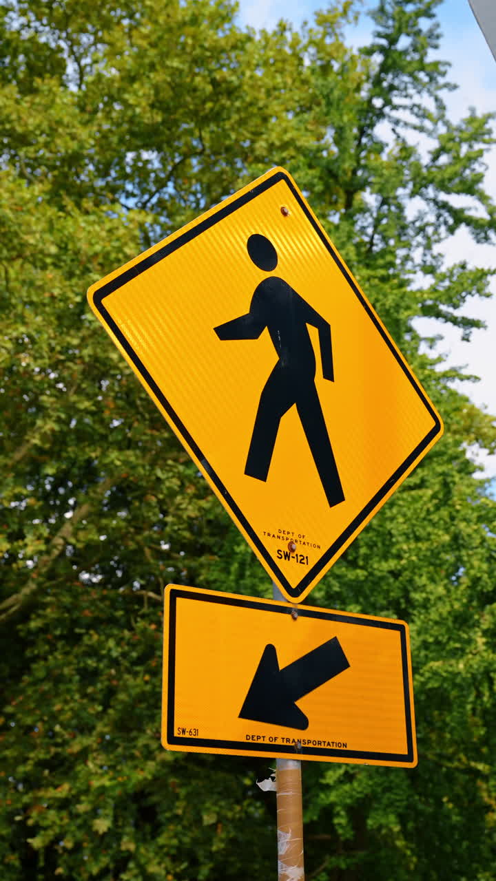 Caution sign for pedestrian zone in park. A bright yellow caution sign warns of pedestrians in a park, with trees behind and an arrow pointing left