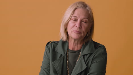 Blonde-Mature-Woman-Wearing-Green-Jacket-Looking-At-Side-Posing-On-Orange-Background,-Then-Touchs-Her-Chin-And-Her-Hair-And-Looks-At-Camera