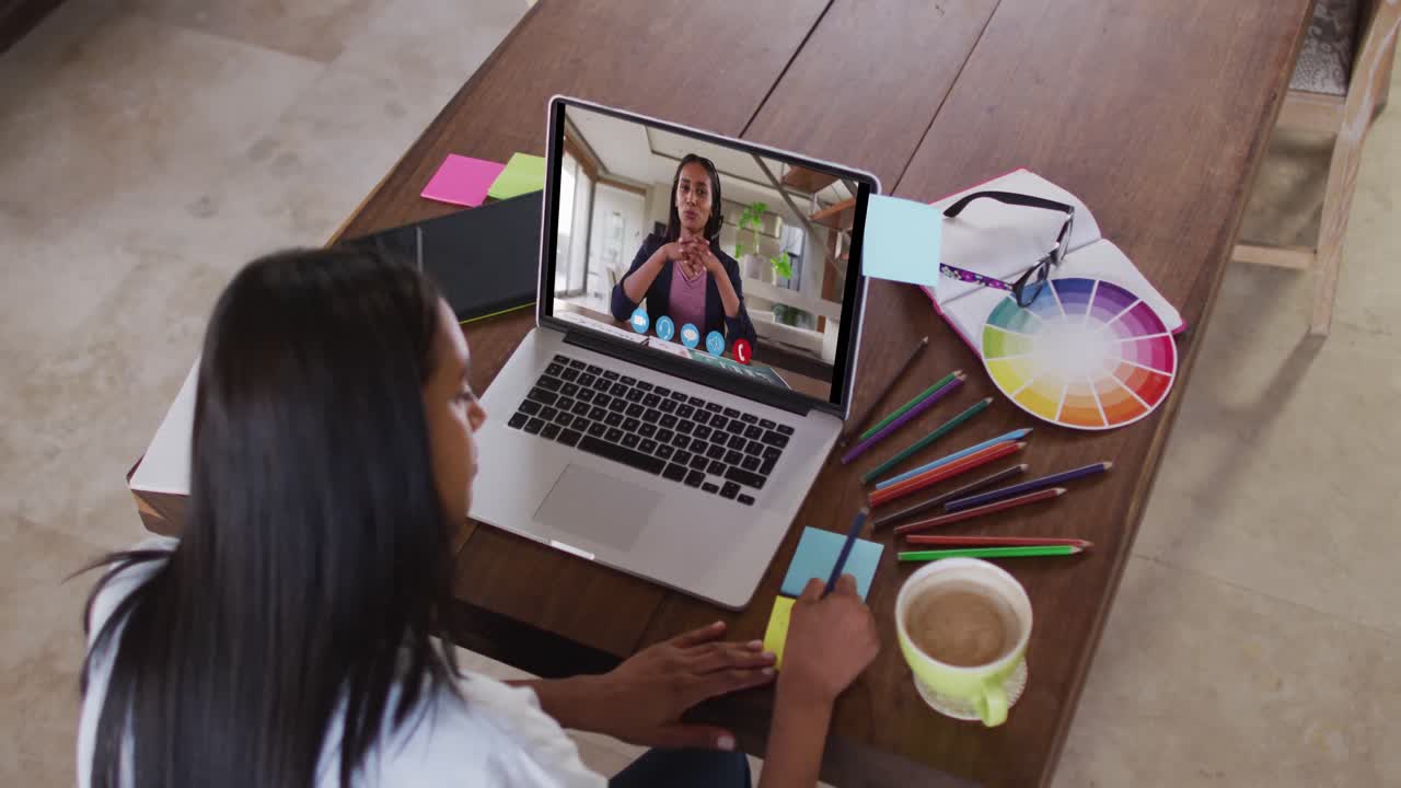 Mujer caucásica usando una laptop en videollamada con una colega femenina, tomando notas y trabajando desde casa