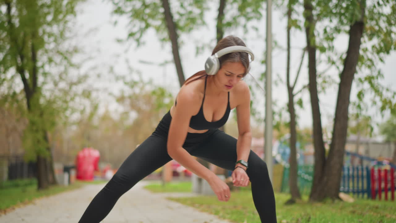 Beautiful athlete stretching her legs while listening to music with headphones, wearing black leggings and top, outdoor park setting, focusing on health, fitness, and stretching