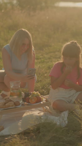 madre e hija sentadas juntas al aire libre haciendo picnic en un día soleado, madre usando teléfono inteligente mientras la hija come frutas, disfrutando del tiempo juntos en la alfombra de picnic con una variedad de bocadillos
