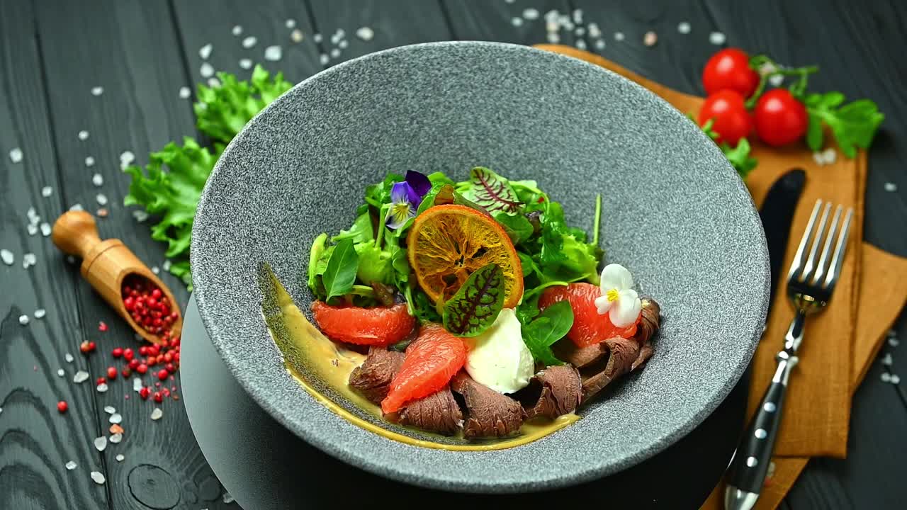 Insalata fresca con carne e agrumi su un tavolo di legno scuro. Un'insalata con carne, agrumi e verdure è disposta su un tavolo di legno nero con utensili da cucina nelle vicinanze.