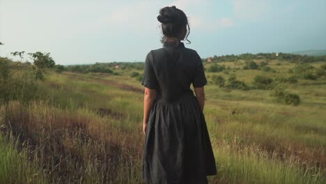 Brunette-woman-in-black-dress-standing-on-a-green-meadow-and-blue-sky-background