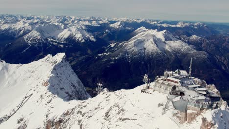 Antena-De-Un-Edificio-En-La-Cima-De-Un-Glaciar-Rodeado-De-Montañas-Nevadas-Y-Un-Valle-Verde