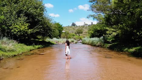 amplia toma lenta de una mujer en un vestido blanco caminando felizmente a través de un arroyo de agua dulce en el medio del bosque