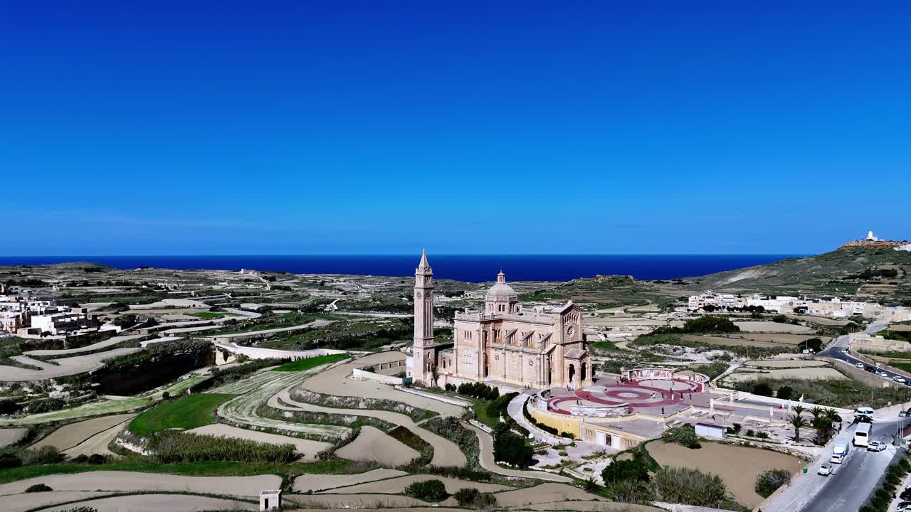 Maestosa vista aerea della Basilica di Ta' Pinu a Gozo, Malta, durante una soleggiata giornata estiva, con architettura storica in pietra calcarea tra campi terrazzati verdi sotto un cielo azzurro limpido e l'orizzonte mediterraneo