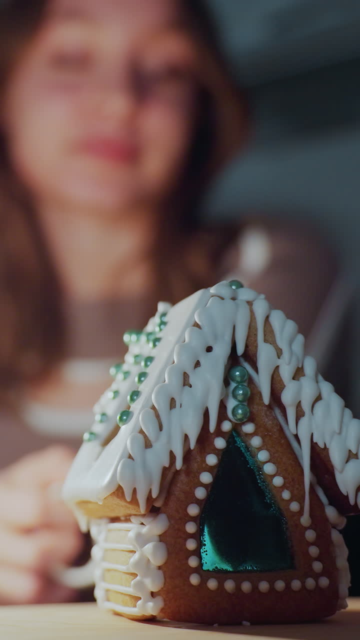 primer plano de una vela roja ardiendo al lado de una casa de pan de jengibre detallada con glaseado blanco, la mujer en el fondo bebe té, ligeramente borrosa, con una iluminación suave y cálida