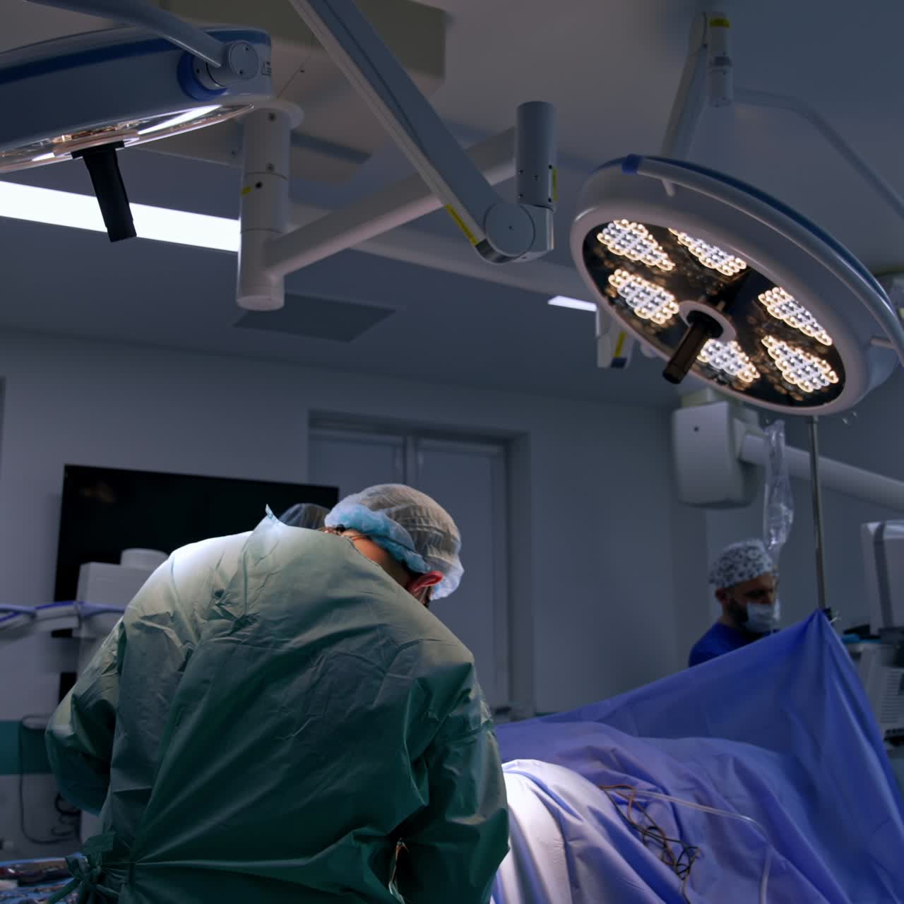 Rear view of surgeon who bent above the operated patient. Female nurse stand opposite to the doctor assisting