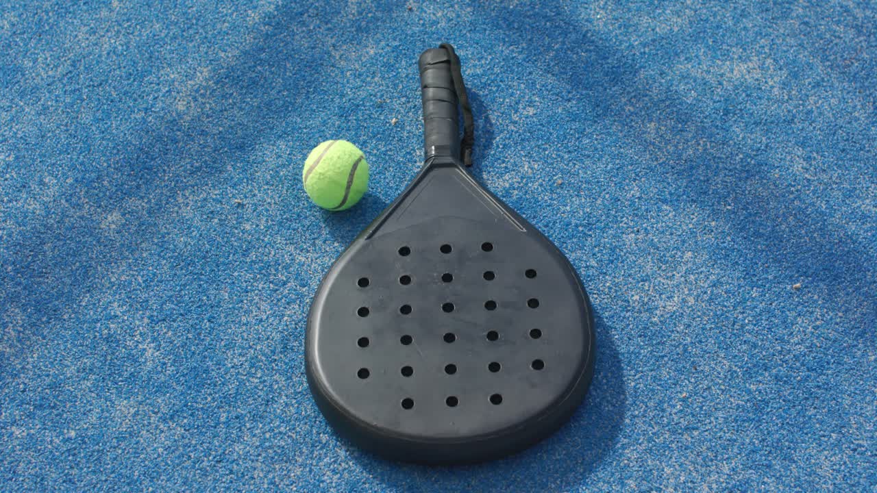 Eine Kamerabewegung schiebt einen Ball um ein Padel-Paddel auf einem blauen Platz, wobei sich der Ball nahe des Schlägerherzens ablegt.