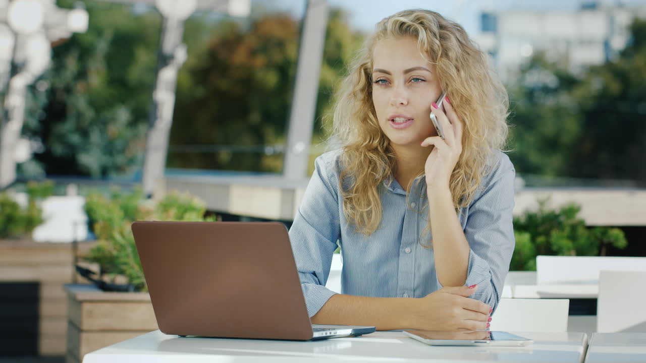 Affascinante donna d'affari che parla al telefono seduta in un caffè sulla terrazza, mentre lavora con un laptop.