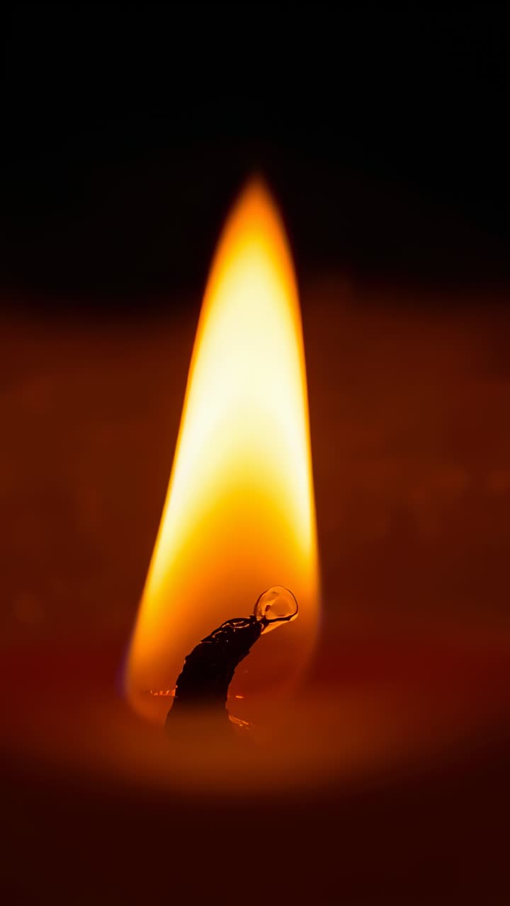 Vertical video: Flickering yellow-orange flame making droplet quiver on candle wick in interior