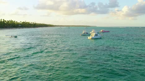 Scenic-flight-above-green-ocean-sea-towards-motorboats-moored-in-water-at-sunset,-overhead-aerial-approach