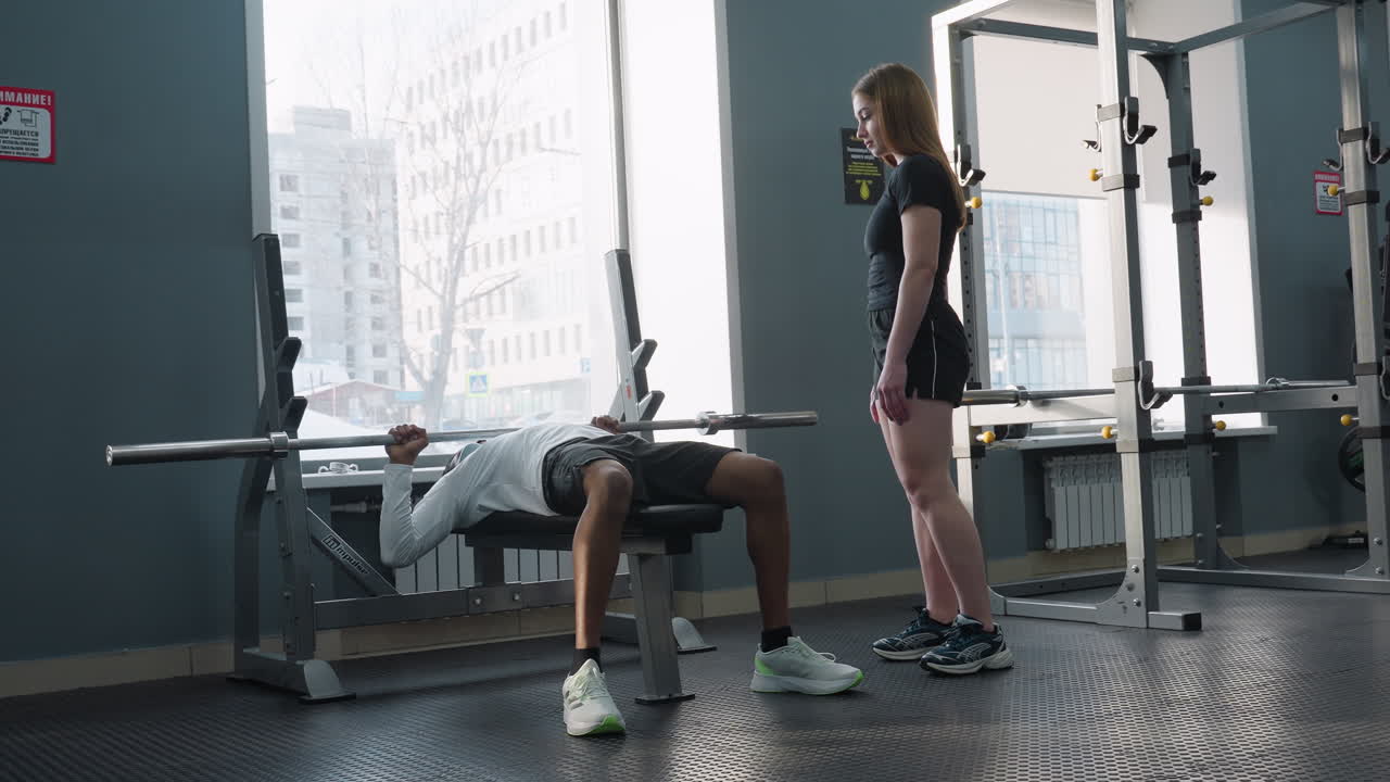 young woman watches friend lift barbell on flat bench inside modern gym with window city view passing car people outside while observing workout session