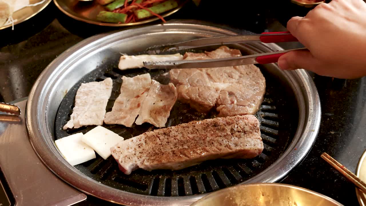 Hand uses tongs to flip pork belly slices on tabletop grill under warm, ambient lighting