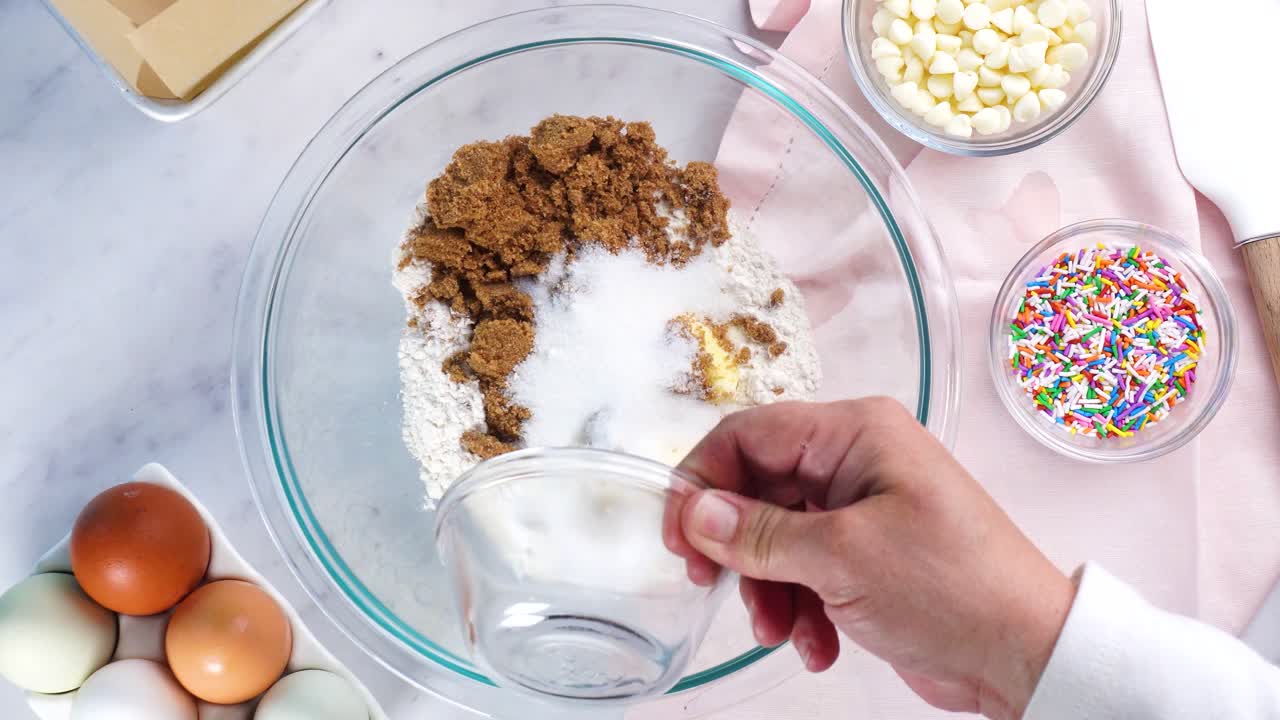 Baking confetti sugar cookies and using a white electric mixer and adding sugar to the batter. In the kitchen making a dessert. Making confetti cookies with a white background, pink napkin and eggs