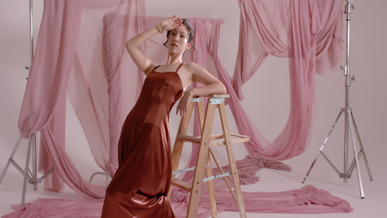 Asian model responding to camera focus in studio, rising onto wood ladder and smoothing satin dress