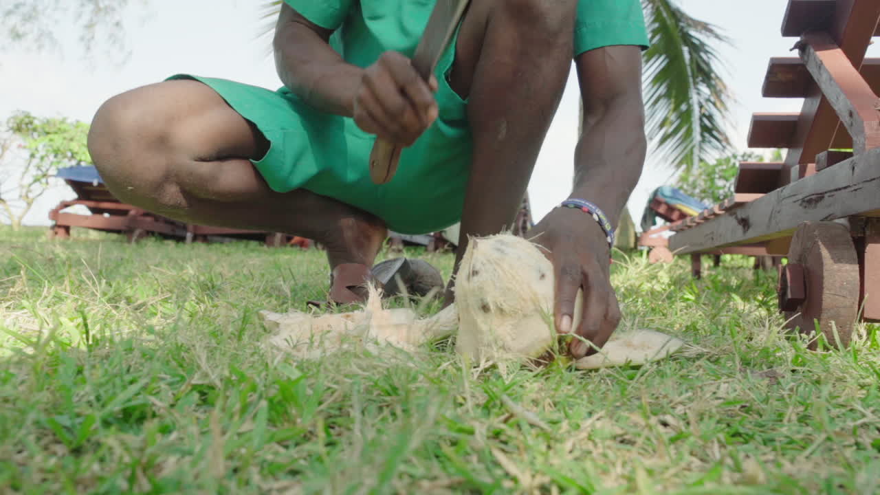 Slow motion footage of island hotel employee opening coconut with machete and knife. Black man cuts coconut open with long knife.