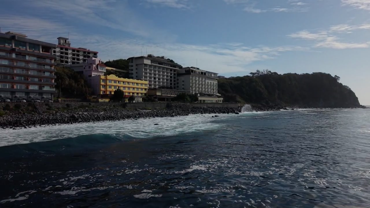Edifici alberghieri a più piani sopra una costa rocciosa alla base di un promontorio boscoso a Higashiizu, Izu, Giappone, illuminati dal sole mattutino velato, ripresa aerea con drone.