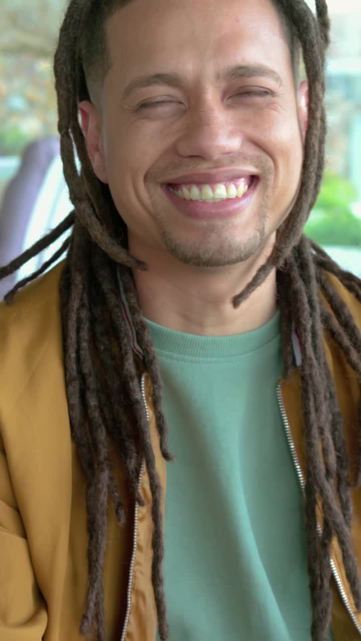 Video verticale: Uomo sorridente con i dreadlock in giacca color cachi che reagisce a una battuta e socchiude gli occhi sul divano