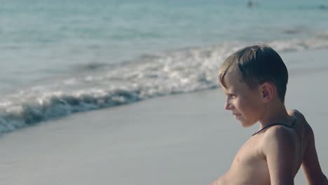 Un-Niño-Feliz-Se-Sienta-Sonriendo-En-La-Línea-De-Surf-Del-Océano-De-Una-Playa-Tropical