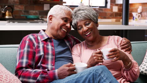 Una-Pareja-De-Ancianos-Sonriente-Relajándose-Con-Una-Bebida-Caliente-En-El-Sofá-De-Casa