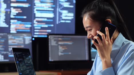 close up side view of asian female programmer talking on smartphone while writing code by a laptop using multiple monitors showing database on terminal window desktops in the office