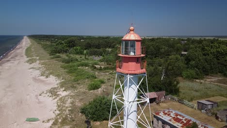 beautiful aerial view of white painted steel lighthouse with red top located in pape, latvia at baltic sea coastline in sunny summer day, wide angle ascending drone shot moving forward close