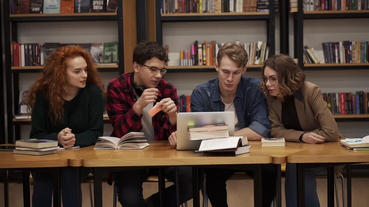 Group of young students talking using laptop explaining course work study together in library. Actively discussing something sitting by wooden table. Slow motion. Front view