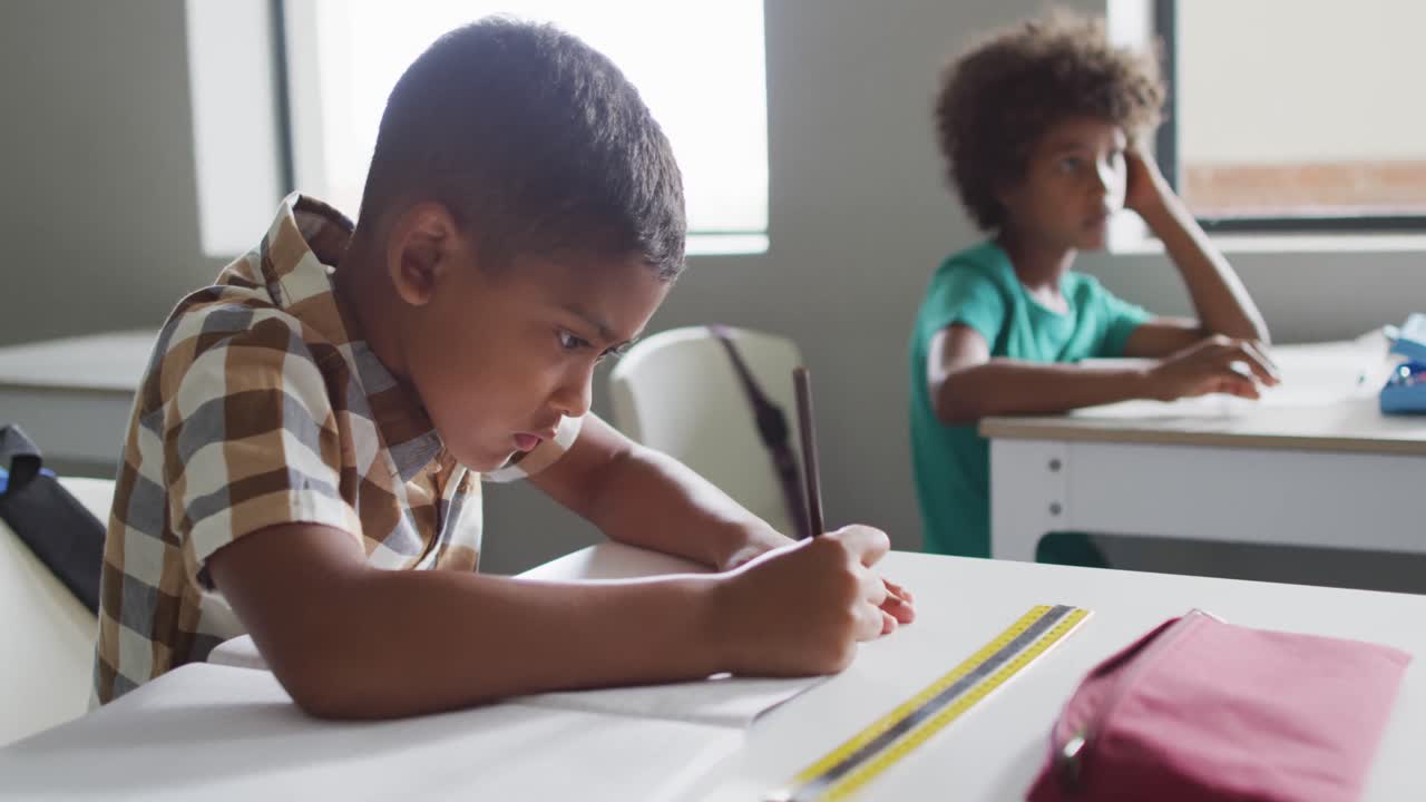 Video of focused biracial boy sitting at desk in classroom
