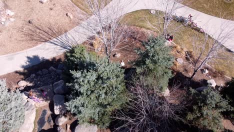 Drone-flying-over-public-rock-park-as-kids-play-and-run,-panning-up-to-reveal-parking-lot,-neighborhood,-and-beautiful-snow-capped-mountains