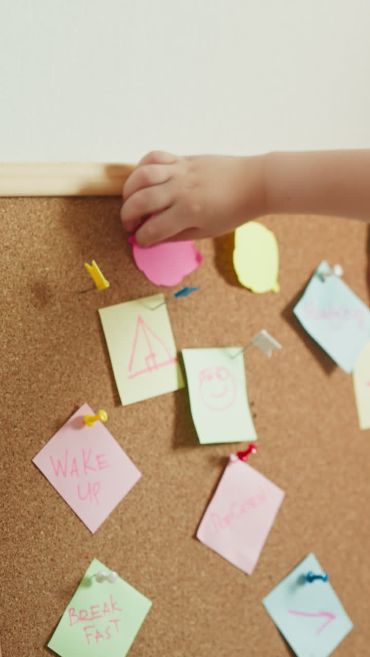 Toddler boy presses pink paper figure to cork board slow motion. Blond kid rests playing with bright colorful notes backside view. Education of children closeup