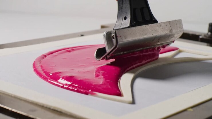 Pulling metal squeegee, operator dragging glossy magenta ink on studio platen, filling paper cutout