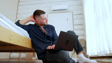thoughtful bearded male sitting on the floor near the bed looks at window. low angle view at the man holding a laptop working at home.