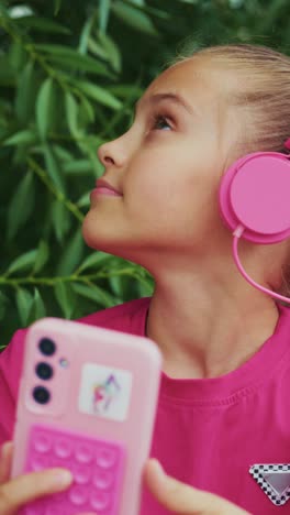 a young girl enjoying music with pink headphones while engaged with her phone surrounded by lush greenery, radiating joy and positivity