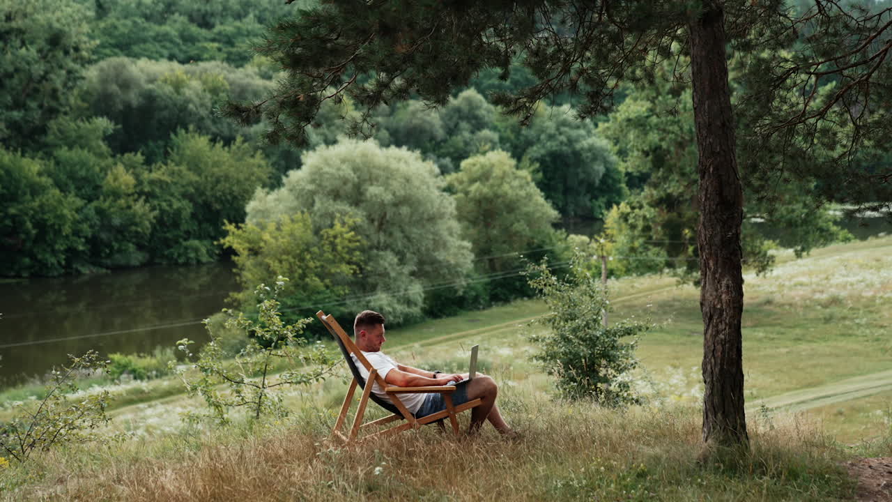 Man works in a calm place among the nature. Entrepreneur types on laptop sitting in a chair.