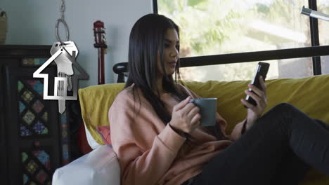 Animación-De-La-Llave-De-La-Casa-Plateada-Y-El-Llavero-Sobre-Una-Mujer-Birracial-Feliz-Usando-Un-Teléfono-Inteligente-En-Casa