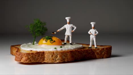 two miniature chefs are standing on a slice of toasted bread with a fried egg, one chef is adding spices to the yolk and the other is watching, isolated on a white background