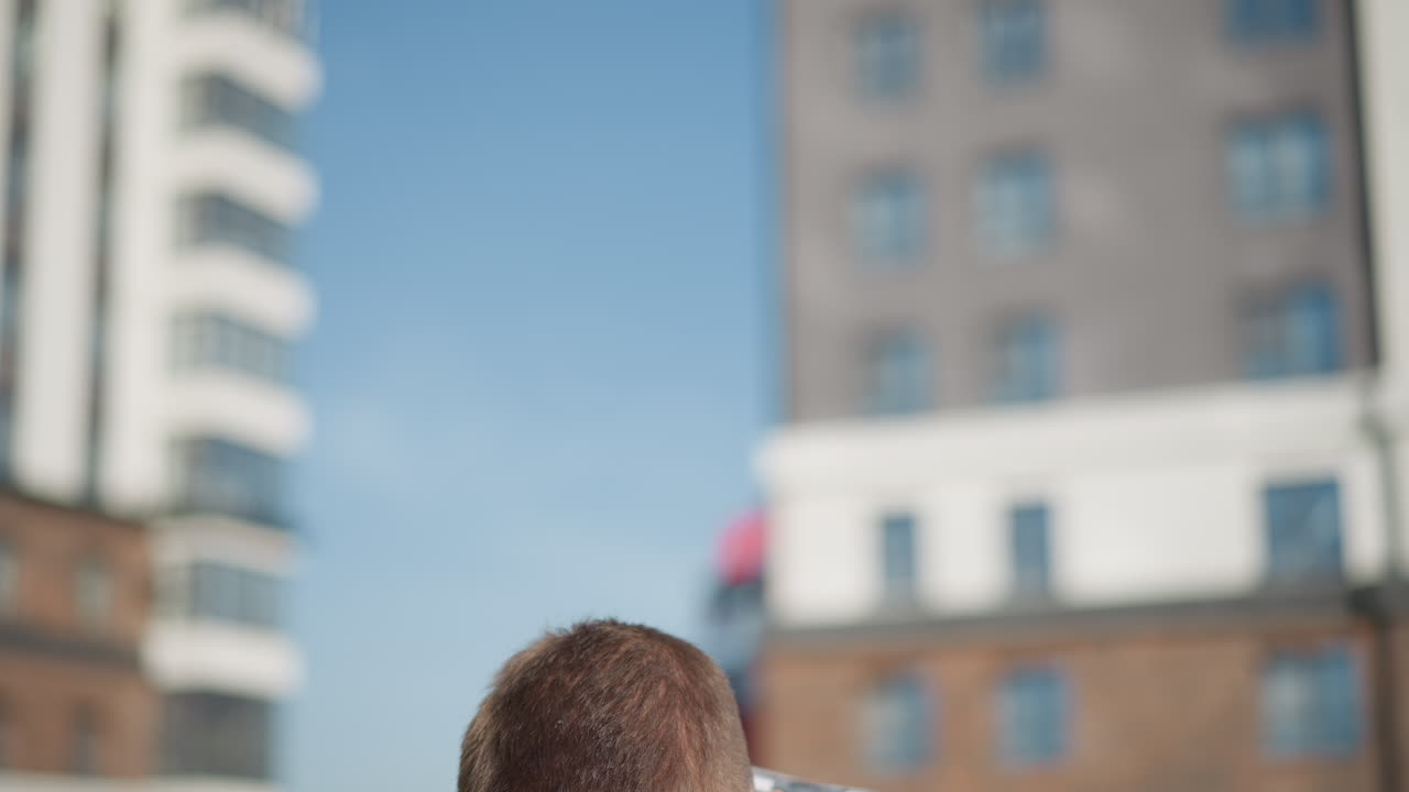 boy wearing checkered shirt sits under bright sun pulls pill from front pocket during calm moment with soft blur of tall urban buildings and modern structures