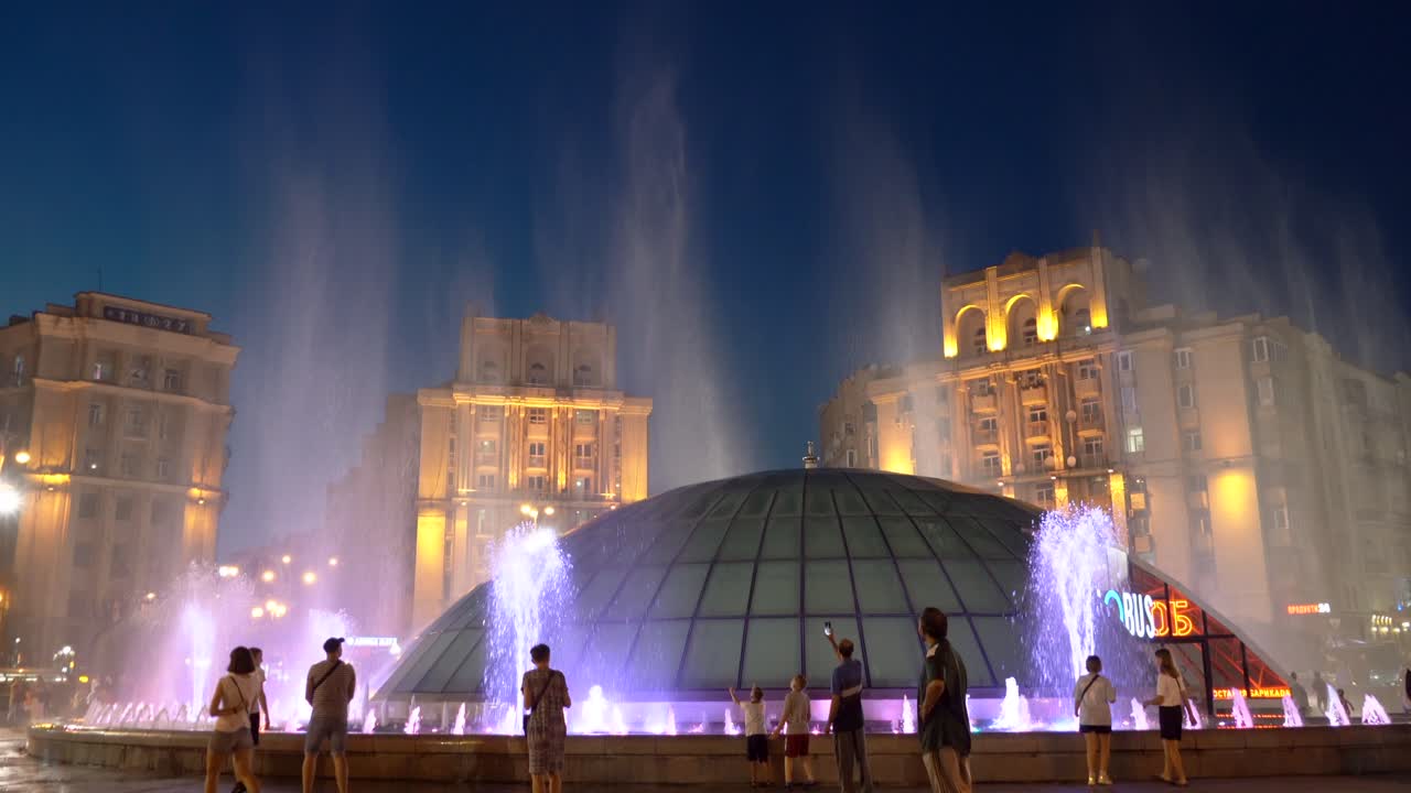 Ukraine, Kyiv, Independence Square at night time, beautifully lighted, flushing fountain in colors with glass dome and people walking around it.