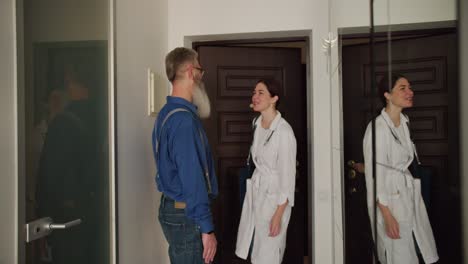 un hombre con cabello gris y una larga barba gruesa en una camisa azul conoce a una chica médica que vino a su casa para un examen en el hogar. una chica con un abrigo blanco de médico viene a un hombre en una visita a casa