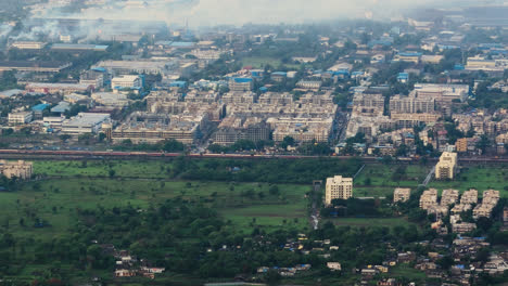 drone moves from left to right over a cityscape, indian railway passing through, surrounded by buildings and lush greenery on both sides with foggy weather above at mumbai