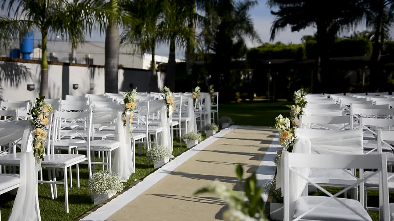 lugar al aire libre decorado para una ceremonia de boda, con sillas blancas colocadas sobre hierba verde, moqueta en el pasillo central decorada con flores y velos, vista desde el altar