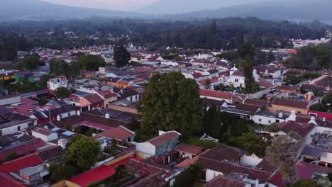gran tiro aéreo de un árbol enorme que se eleva a través de los tejados al amanecer en antigua, guatemala, con los volcanes acatenango y fuego visibles
