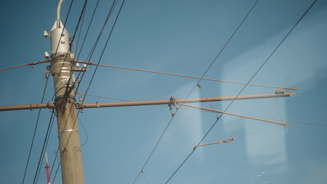 vehicle moves past city utility pole with overhead wires against clear blue sky, view through window shows light reflections as street infrastructure slides by, motion suggests transit commute
