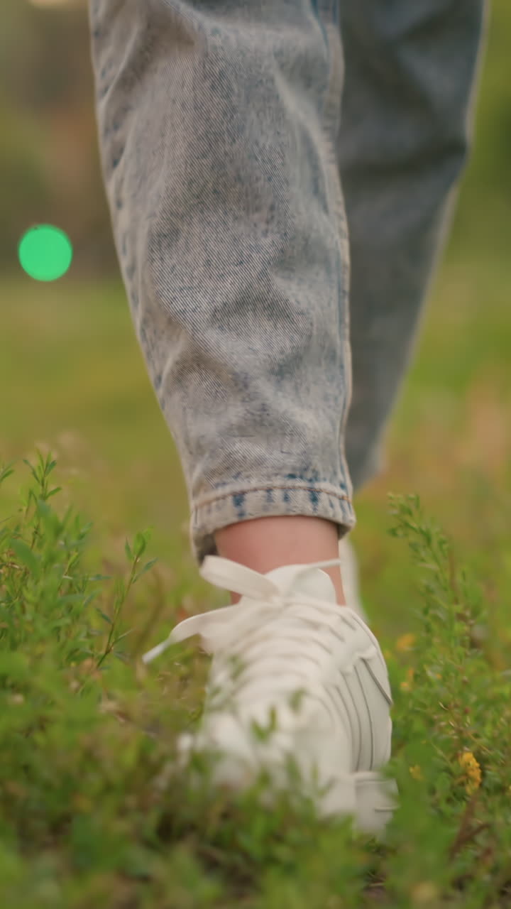 Close-up of legs in white sneakers and rolled-up jeans, walking through a grassy field with patches of brown foliage, blurred red and green lights appear in the distant background