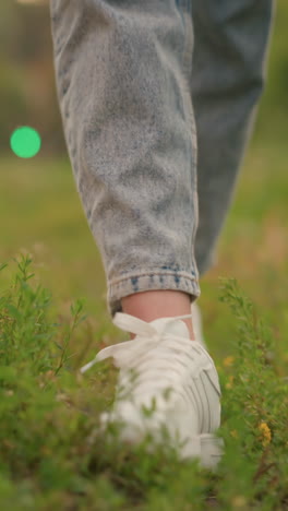 primer plano de piernas en zapatillas blancas y vaqueros enrollados, caminando a través de un campo cubierto de hierba con manchas de follaje marrón, luces rojas y verdes borrosas aparecen en el fondo lejano