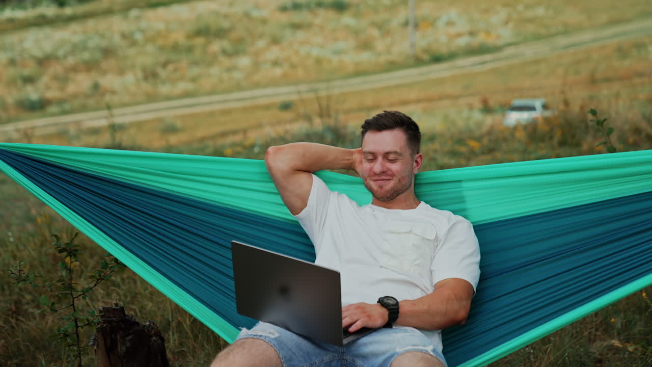 Joyful Caucasian man sits in hammock with laptop on his knees. Relaxed man working distantly away from office.