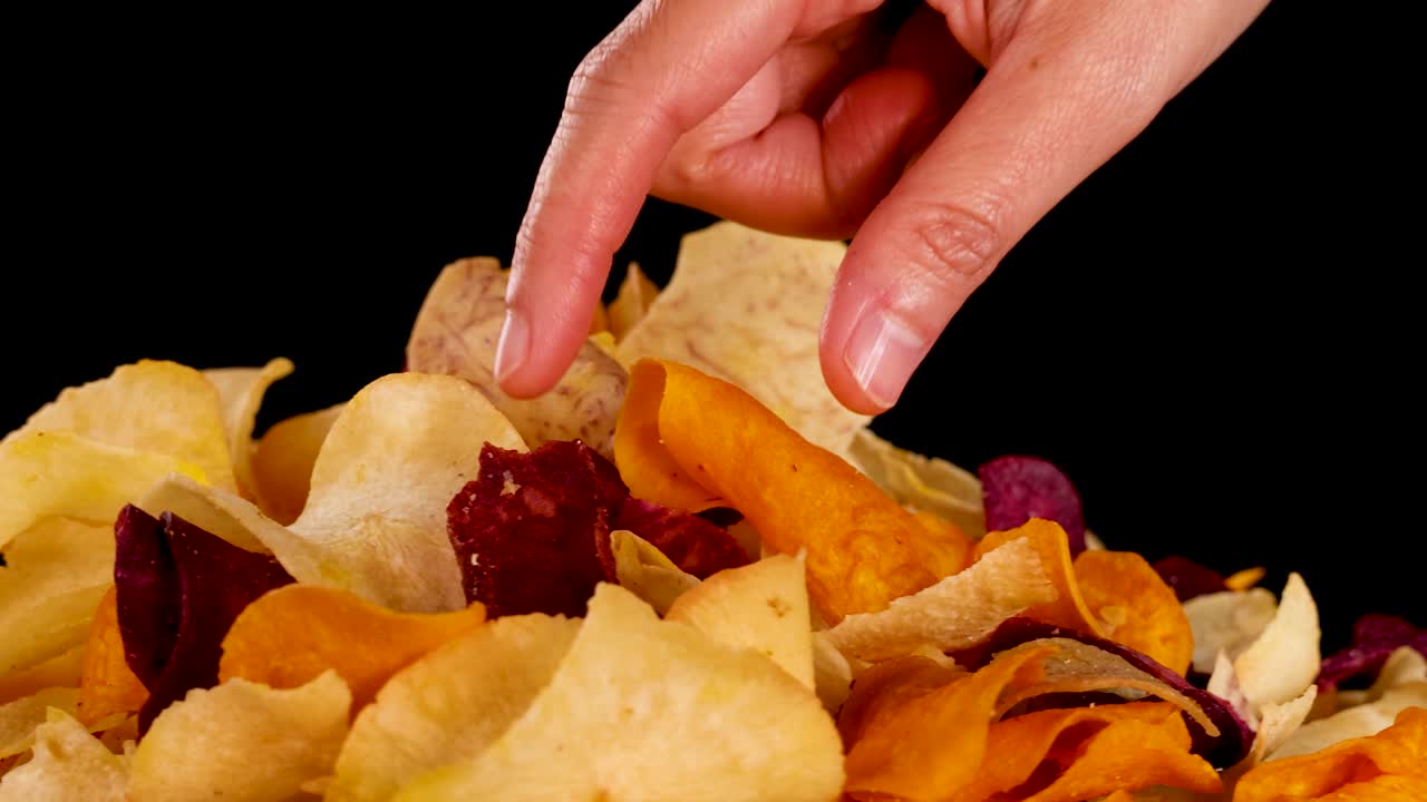 Una mano raccoglie chips di colorate verdure a radice sotto luci luminose da studio, primo piano, sfondo nero