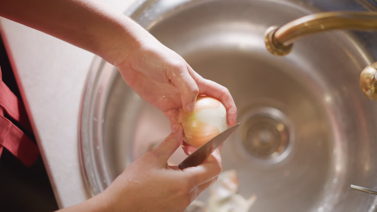 Hands peeling potato over sink with peeler and knife, peels falling into basin beneath brass faucet, focused home cook rhythm and practical technique, towel and apron hint, tactile motion and textured
